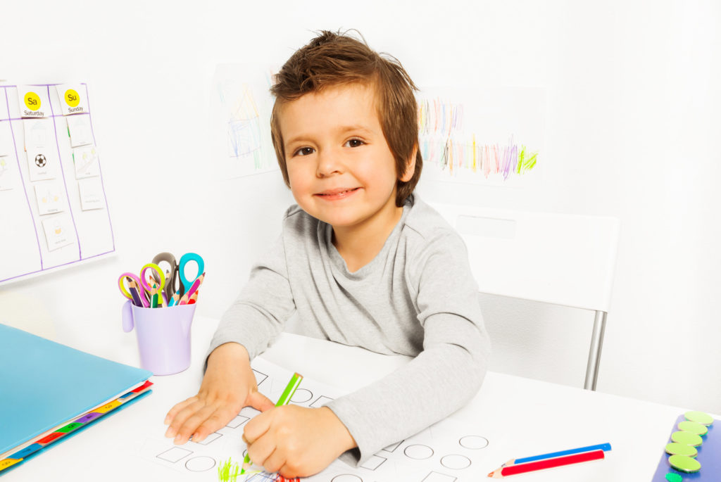 Positive small boy draws with pencil during Applied Behavior Analysis ABA sitting at the table indoors